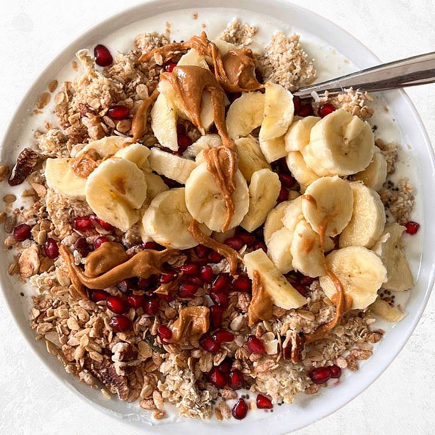 Bowl con porridge, banana, granola Clemi's Market, melograno e burro di arachidi con cucchiaio argento